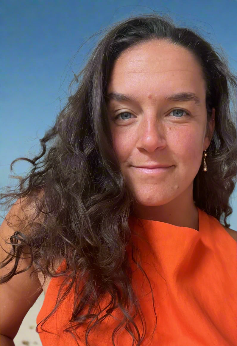 Woman with long dark hair wearing an orange top in a room with a bookshelf.